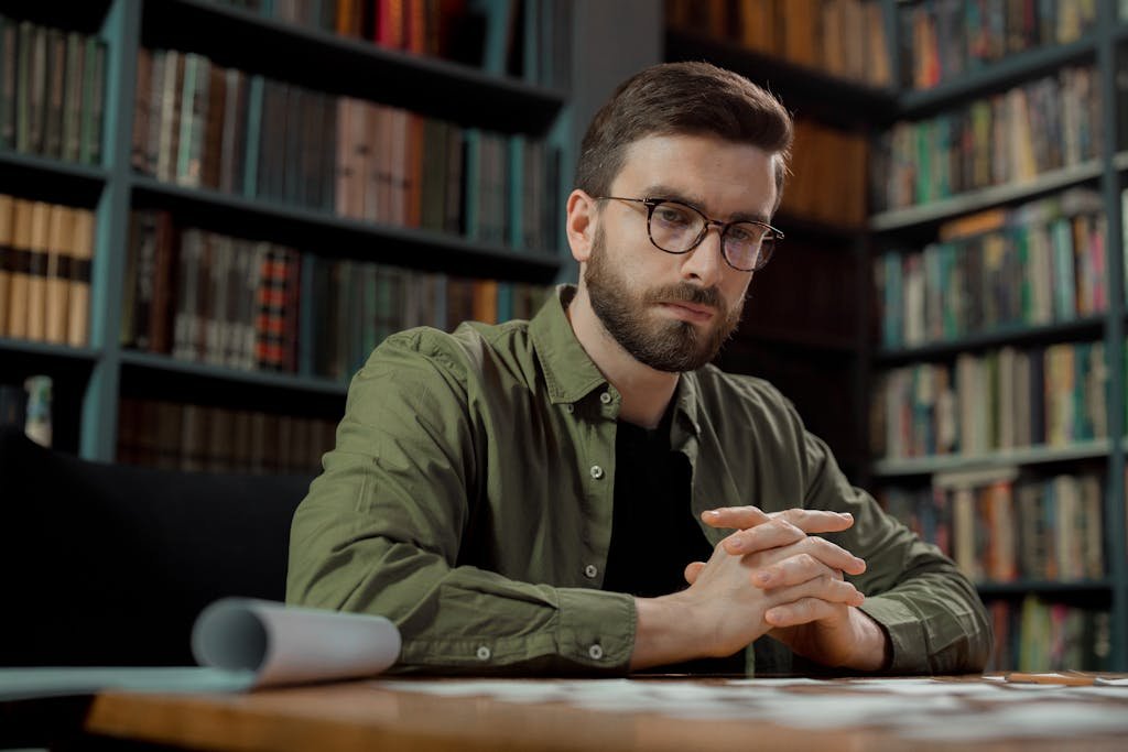 A contemplative male adult seated in a library, wearing eyeglasses and deep in thought.